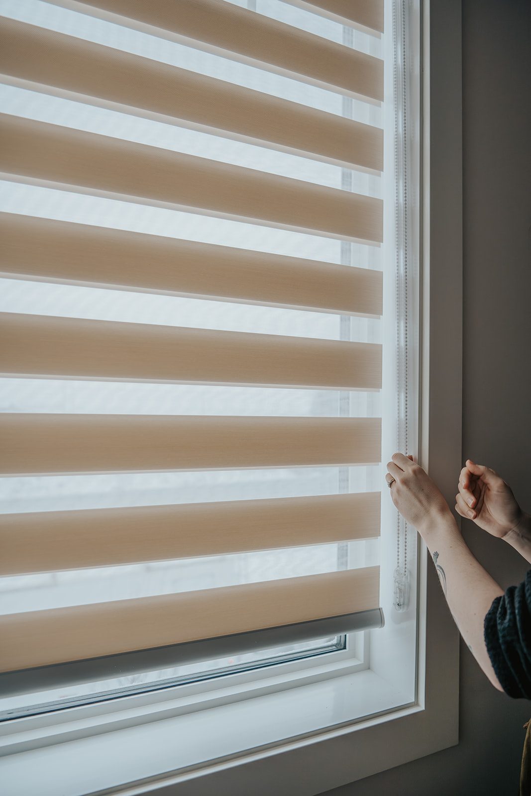 A hand adjusting a Zebra blind beside a window.
